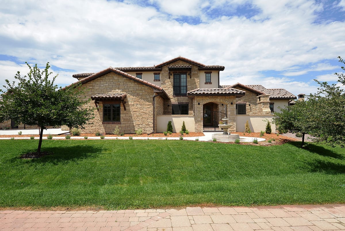 A Tuscan-style stone home with a tile roof behind a lawn